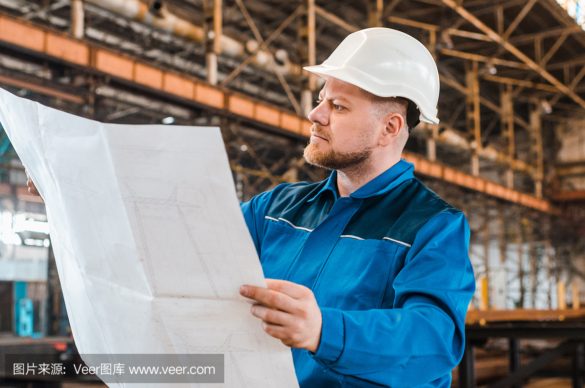 一個(gè)穿著工作服、戴著白色頭盔的工人正在檢查一個(gè)金屬工廠背景上的圖紙。建筑工人身體很好。男工程師